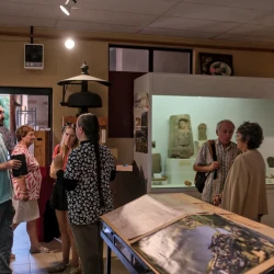Vue large d'une grande pièce avec des vitrines, des photos, et des personnes qui regardent et échangent. En arrière plan à gauche, une vue de l'extérieur ensoleillé.