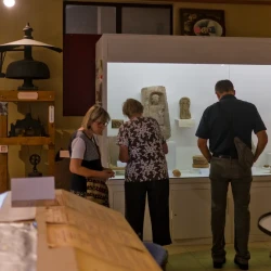 Vue large d'une grande pièce avec des vitrines, des photos, et des personnes qui regardent et échangent. En arrière plan à gauche, une vue de l'extérieur ensoleillé.