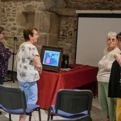 Dans une pièce aux murs en pierre, 4 femmes échangent des regards devant un écran dans un cadre de tableau qui monter une photo d'un couple.