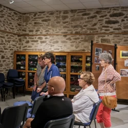 Dans une pièce aux murs en pierre, des personnes se tenant debout et assises regardent toutes vers la gauche. Au fond, une vitrine avec des maquettes de camions, et à droite des photos anciennes sur des panneaux en bois.