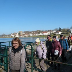 Randonneurs sur un barrage, devant un étang et un village, sous un ciel bleu
