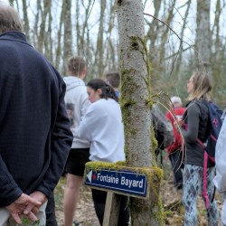Des marcheurs en pause, vue de dos, et au centre une pancarte moussue et adossée à un petit tronc d&#039;arbre qui indique Fontaine Bayard avec une flèche sur la gauche.
