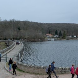 Marcheurs devant et sur un barrage, avec le plan d&#039;eau sur la droite. Au loin le reste des marcheurs sur une route, devant une forêt.