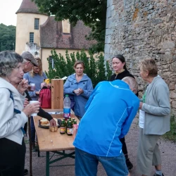 Des personnes autour d'une table devant un mur de pierre et des arbres avec des verres à la main et qui grignotent.