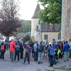 Un groupe de personnes rassemblées en train de se restaurer et boire, devant un édifice avec une tour, 1 arbre. À droite, on distingue un mur en pierre d&#039;un bâtiment ancien.