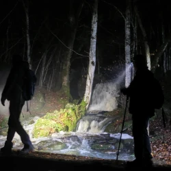 Au 1er plan, des marcheurs sur un passage au-dessus d'un ruisseau, à l'arrière plan une cascade au milieu des bois éclairée par des lampes de marcheurs.