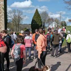 Des randonneurs rassemblés sur une place, on distingue l'angle d'un grand mur en pierre à gauche, quelques arbres et maisons en arrière plan sous un ciel bleu avec quelques nuages