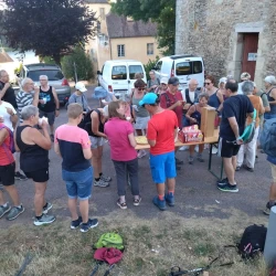 Un groupe de marcheurs sur une esplanade à l'ombre, en train de boire un coup. À droite une vieille tour en pierre, au centre des arbres, au pied, des voitures et plus loin un autre bâtiment ancien, des maisons, et un lac et une forêt en arrière plan.