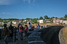Un groupe de marcheurs sur un barrage : la partie la plus proche est à l'ombre, le reste au soleil. Au loin on aperçoit un village.