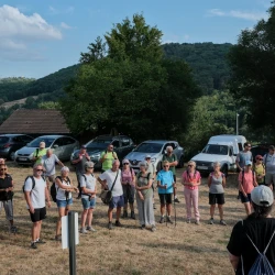 Des gens sur un parking en herbe entourés de voitures écoutent 2 personnes en train de s'adresser à elleux. Au loin de la forêt vallonnée et un ciel bleu et nuages.