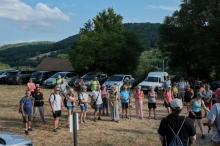 Des gens sur un parking en herbe entourés de voitures écoutent 2 personnes en train de s'adresser à elleux. Au loin de la forêt vallonnée et un ciel bleu et nuages.