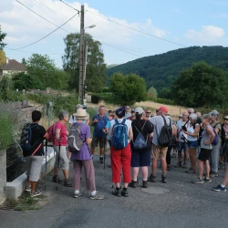 Un groupe de marcheurs est arrêté près d'une fontaine qu'on distingue sur la gauche derrière de la lavande, et écoute les explications du guide. Des maisons disséminées autour, et une colline en forêt en arrière plan sous un ciel bleu avec nuages.