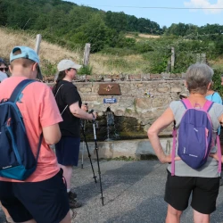 Quelques marcheurs arrêtés près d'une fontaine qu'on voit de face. Derrière, un pré et de la forêt.