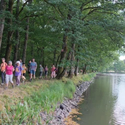 À gauche, des marcheurs en file indienne sur un chemin bordé d'arbres le long d'un étang sur la droite.