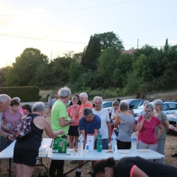 Des gens autour de tables pliantes en train de boire. Derrière des voitures, plus loin des maisons entourées de végétation.