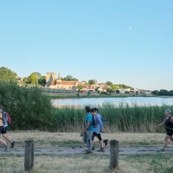 7 marcheureuses sur un chemin devant un étang et sa végétation. En arrière plan, un bourg de village où on voit une église, une tour carrée, un cimetière et des bâtiments divers, sous le soleil rasant du soir. Au 1er plan, trois potelets en bois.