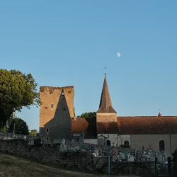 Au premier plan un cimetière et son mur d'enceinte à l'ombre. Derrière, de gauche à droite, un arbre, une tour carrée et une église, dans les rayons rasants de soleil. L'ombre du clocher se projette sur la tour. La lune au-dessus et légèrement à droite du clocher complète la photo.